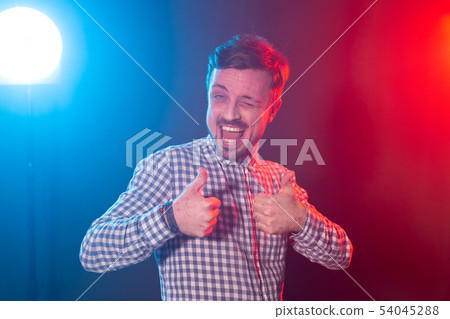 Laughing young bearded man in a plaid shirt showing thumb up posing in studio on blue-red background 54045288