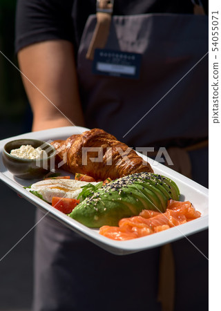 Breakfast is set. Whole grain sandwich with fried egg, red fish, avocado. Plate in the hands of a 54055071