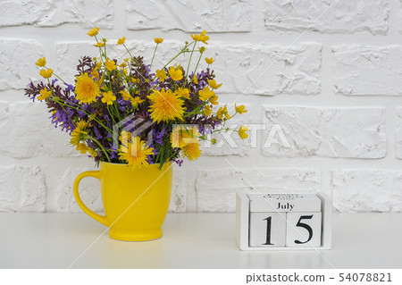 Wooden cubes calendar July 15 and yellow cup with bright colored flowers against white brick wall Wooden cubes calendar July 15 and yellow cup with bright colored flowers against white brick wall 54078821