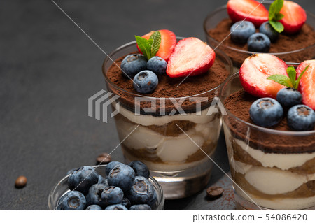 Classic tiramisu dessert with blueberries and strawberries in a glass on stone serving board on dark Classic tiramisu dessert with blueberries and strawberries in a glass on stone serving board on dark 54086420