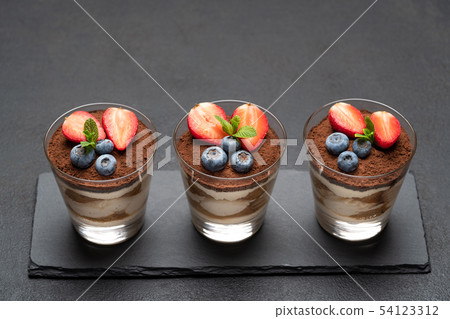 Classic tiramisu dessert with blueberries and strawberries in a glass on stone serving board on dark Classic tiramisu dessert with blueberries and strawberries in a glass on stone serving board on dark 54123312