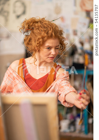 Curly artist looking at painting brush while working Curly artist looking at painting brush while working 54135707