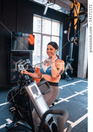 Dark-haired woman cycling in gym and speaking on phone Dark-haired woman cycling in gym and speaking on phone 54171172