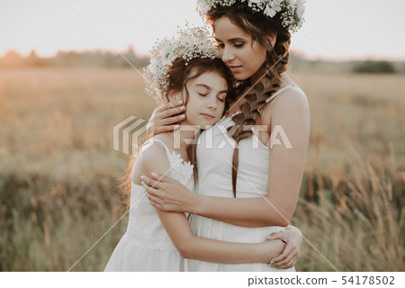 mother and daughter hugging together in white dresses with braids and floral wreaths in boho style 54178502