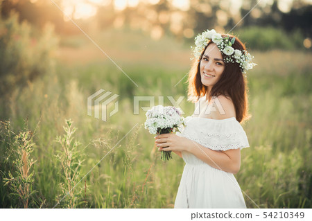 Caucasian happy girl in a white dress with a bouquet of flowers and a wreath in the summer 54210349