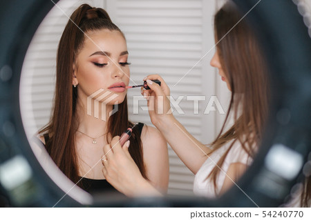 Photo though the ring lamp of makeup artist doing a makeup for attractive young girl. Background of 54240770