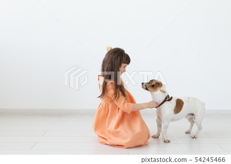 Little girl with dark hair plays with her beloved dog while sitting on the floor in a peach dress 54245466