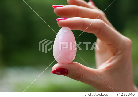 Close up photo of female hand holding a vaginal yoni egg. Rose quartz crystal egg on river Close up photo of female hand holding a vaginal yoni egg. Rose quartz crystal egg on river 54253340