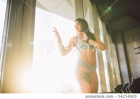 Young caucasian sporty woman with curly long hair poses in sportswear near a panorman window in a Young caucasian sporty woman with curly long hair poses in sportswear near a panorman window in a 54254920
