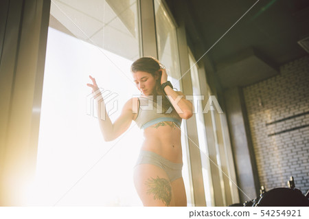 Young caucasian sporty woman with curly long hair poses in sportswear near a panorman window in a Young caucasian sporty woman with curly long hair poses in sportswear near a panorman window in a 54254921