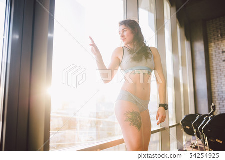 Young caucasian sporty woman with curly long hair poses in sportswear near a panorman window in a 54254925