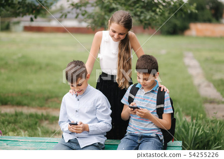 Two boys and girl use their phones during school breack. Cute boys sitting on the bench and play 54255226
