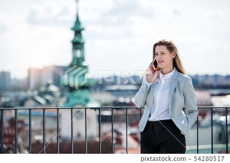A young businesswoman with smartphone standing on a terrace, working. 54280517