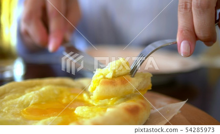 close-up female hands with a knife and fork cut off a slice from Adjara khachapuri with an egg on a close-up female hands with a knife and fork cut off a slice from Adjara khachapuri with an egg on a 54285973