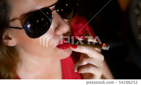 gorgeous woman in red dress with red lipstick on her lips and dark glasses with a cigar on a dark 54286252