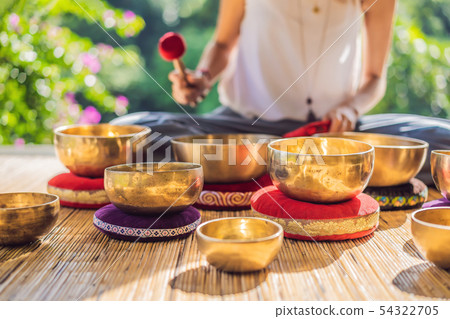 Woman playing on Tibetan singing bowl while sitting on yoga mat against a waterfall. Vintage tonned 54322705