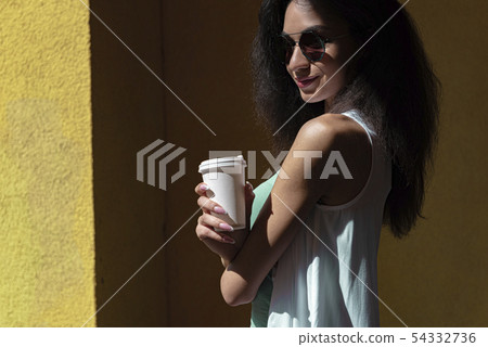 Charming young woman with paper coffee cup standing against yellow wall 54332736
