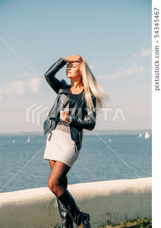 Beautiful blonde in a leather black jacket posing on the shore of the blue sea Beautiful blonde in a leather black jacket posing on the shore of the blue sea 54345467
