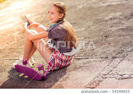 Cheerful long-haired girl staying on the ground with skateboard nearby Cheerful long-haired girl staying on the ground with skateboard nearby 54349855