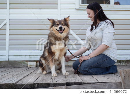 Photo of sitting happy brunette in jeans and white sweater next to dog on leash against background 54366072