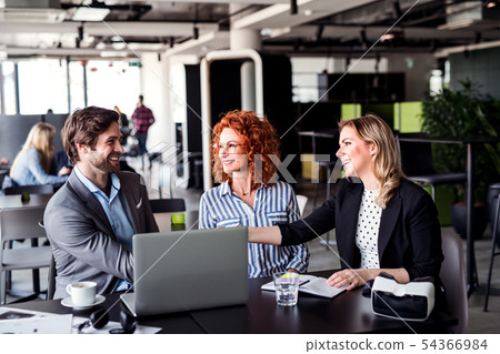 A group of young business people with laptop sitting in an office, talking. A group of young business people with laptop sitting in an office, talking. 54366984