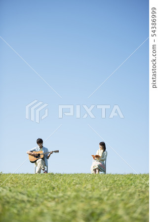 A man reading a book with a man playing guitar on a meadow A man reading a book with a man playing guitar on a meadow 54386999