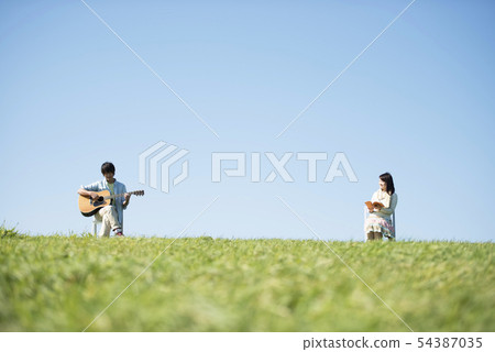 A man reading a book with a man playing guitar on a meadow 54387035