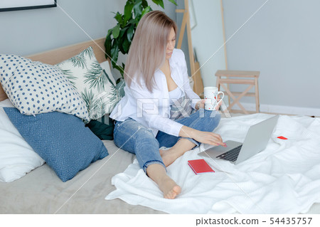 Blonde young woman sits on bed with laptop Blonde young woman sits on bed with laptop 54435757