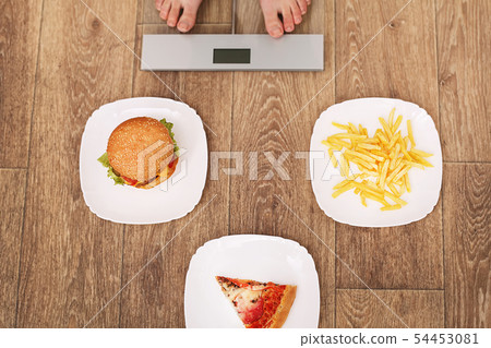 A young beautiful woman is on the scales. To choose between potato chips, pizza and burgers. The 54453081