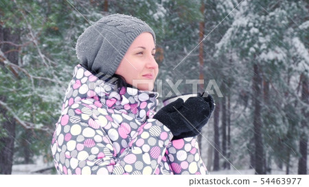 A female tourist stands against a winter forest or park on a snowy day and drinks hot tea from a cup A female tourist stands against a winter forest or park on a snowy day and drinks hot tea from a cup 54463977