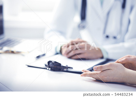 Doctor and female patient sitting at the desk and talking in clinic near window. Medicine and health Doctor and female patient sitting at the desk and talking in clinic near window. Medicine and health 54483234