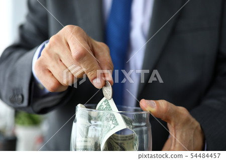Rich man in suit and tie put extra coin into jar full of US dollars Rich man in suit and tie put extra coin into jar full of US dollars 54484457