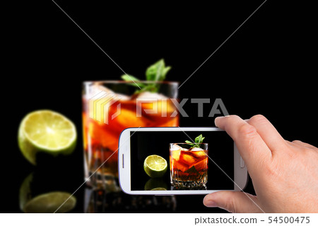 hands of man with smartphone taking photo Glass of Dark Rum Cocktail with lime, orange, ice cubes 54500475