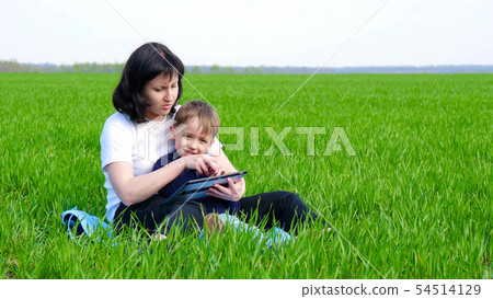 Happy family: mother and child sitting on the green grass. Mom and boy use tablet computer. 54514129