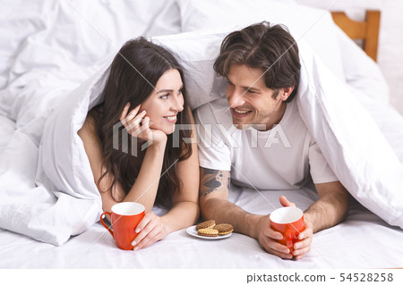 Young couple having breakfast in bed, hiding under blanket Young couple having breakfast in bed, hiding under blanket 54528258
