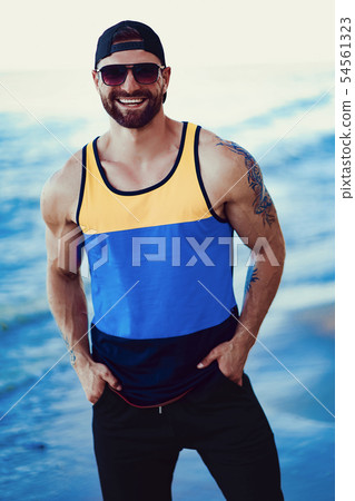 Bearded tattooed man at the resort in a sportive shirt, cap and sunglasses on the sea background Bearded tattooed man at the resort in a sportive shirt, cap and sunglasses on the sea background 54561323