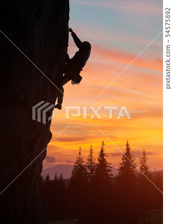 Silhouette climber climbing at nightfall, reaching for next hold hand. Sunset orange cloudy sky. Low 54575892