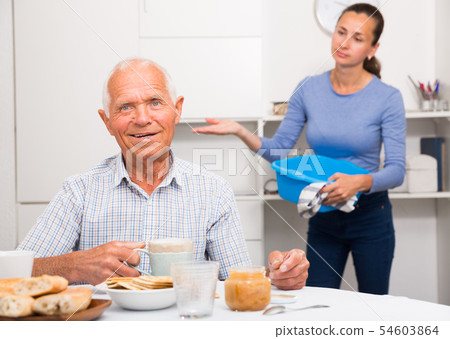 Mature father drinking tea in the kitchen. Adult daughter does cleaning in the apartment Mature father drinking tea in the kitchen. Adult daughter does cleaning in the apartment 54603864