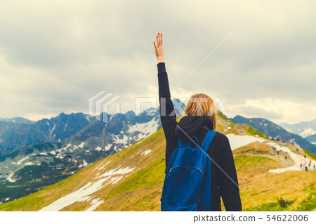 Blonde girl traveler with a blue backpack raised her hand up and enjoying the green mountain scenery Blonde girl traveler with a blue backpack raised her hand up and enjoying the green mountain scenery 54622006