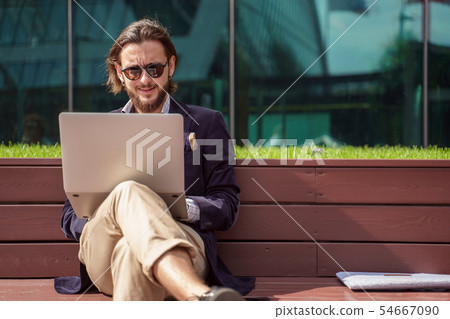 Photo of male freelancer with laptop sitting on wooden bench in center of city on summer day 54667090