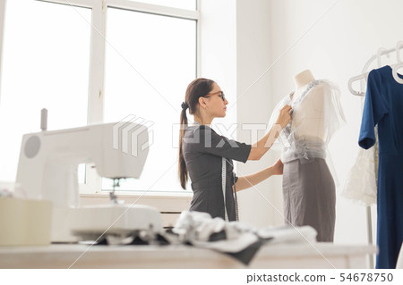 Dressmaker, tailor, fashion and showroom concept - Side view of female fashion designer measuring Dressmaker, tailor, fashion and showroom concept - Side view of female fashion designer measuring 54678750
