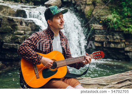 Portrait of a man plays a guitar sitting on a trunk of a tree against a waterfall. Space for your Portrait of a man plays a guitar sitting on a trunk of a tree against a waterfall. Space for your 54702705