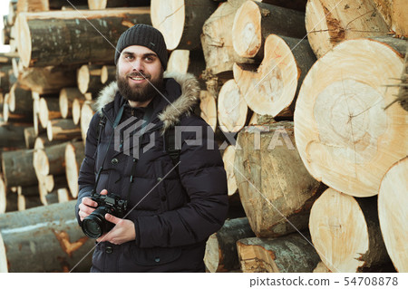 Portrait of a man in the background of a huge pile of fallen trees is holding a camera. Portrait of a man in the background of a huge pile of fallen trees is holding a camera. 54708878