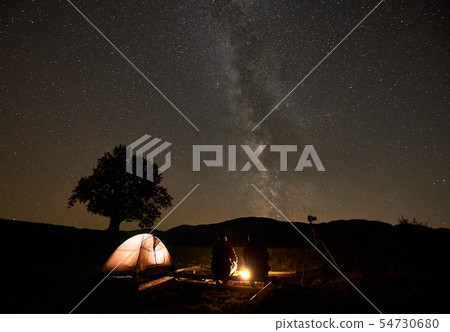 Two tourists at burning campfire in front of tent, photo camera on tripod under dark starry sky. Two tourists at burning campfire in front of tent, photo camera on tripod under dark starry sky. 54730680