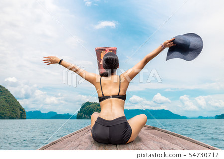 Woman wearing bikini sitting on the boat with hands up and holding beachhat. Woman wearing bikini sitting on the boat with hands up and holding beachhat. 54730712