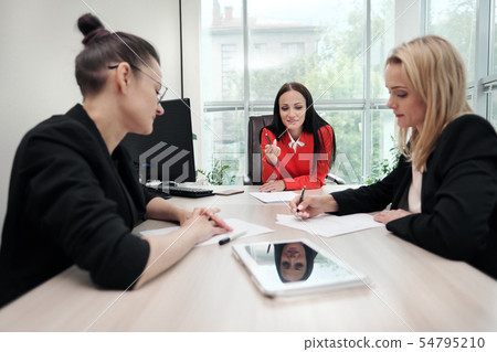 Three young attractive women in business suits are sitting at a desk and discussing workflows. Head Three young attractive women in business suits are sitting at a desk and discussing workflows. Head 54795210