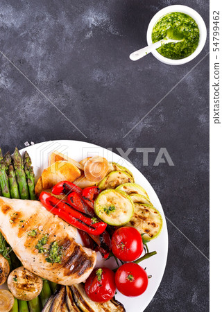 Chicken breast grill with bbq vegetables and pesto sauce in a plate on a concrete background, flat Chicken breast grill with bbq vegetables and pesto sauce in a plate on a concrete background, flat 54799462