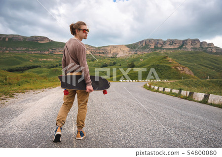 A stylish young man walks along a winding mountain road with a skate or longboard in his hands the 54800080