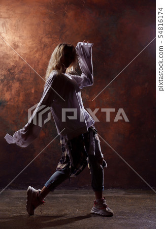Photo of young dancing blonde looking to side in torn jeans and sneakers on dark brown background Photo of young dancing blonde looking to side in torn jeans and sneakers on dark brown background 54816174