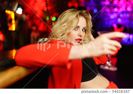 Image of young blonde in red suit looking at camera sitting at bar with cocktail glass in nightclub 54816387